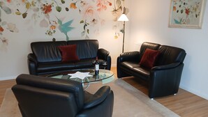 A living room with a glass coffee table, two black leather sofas with red cushions, a floor lamp, and floral wall art. - Ground-Level Holiday Home by the Salt Marshes (Sande)