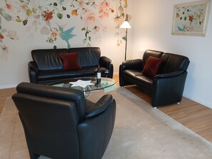 Ein Wohnzimmer mit einem Glastisch, zwei schwarzen Ledersofas mit roten Kissen, einer Stehlampe und floralen Wandbildern.