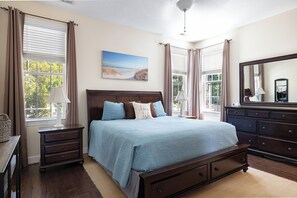 A bedroom with a large bed, a dresser, a nightstand, a lamp, a painting, and a window with curtains. - Blue Byrds - 4 Bedroom Home (Emerald Isle)