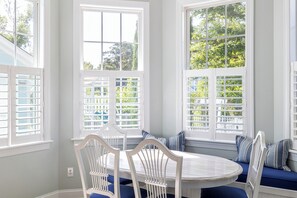 Un coin repas avec une table ronde et des chaises blanches, de grandes fenêtres équipées de volets, et une vue sur la verdure à l’extérieur.