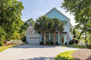 A two-story house with a garage, surrounded by palm trees and a white fence. - Blue Byrds - 4 Bedroom Home (Emerald Isle)