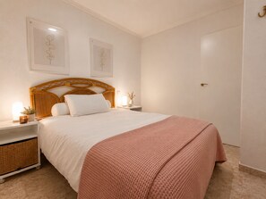Ein ordentlich eingerichtetes Schlafzimmer mit einem hölzernen Kopfteil, einer weißen Bettdecke, einer rosa Strickdecke und zwei an der Wand befestigten Nachttischen mit Lampen und kleinen Pflanzen.