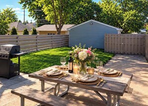 An outdoor dining area with a table set for a meal, a grill, and a house in the background.