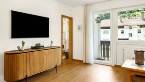 Ein modernes Wohnzimmer mit einem großen Flachbildfernseher, Holzmöbeln und Blick auf Gebäude durch ein großes Fenster.