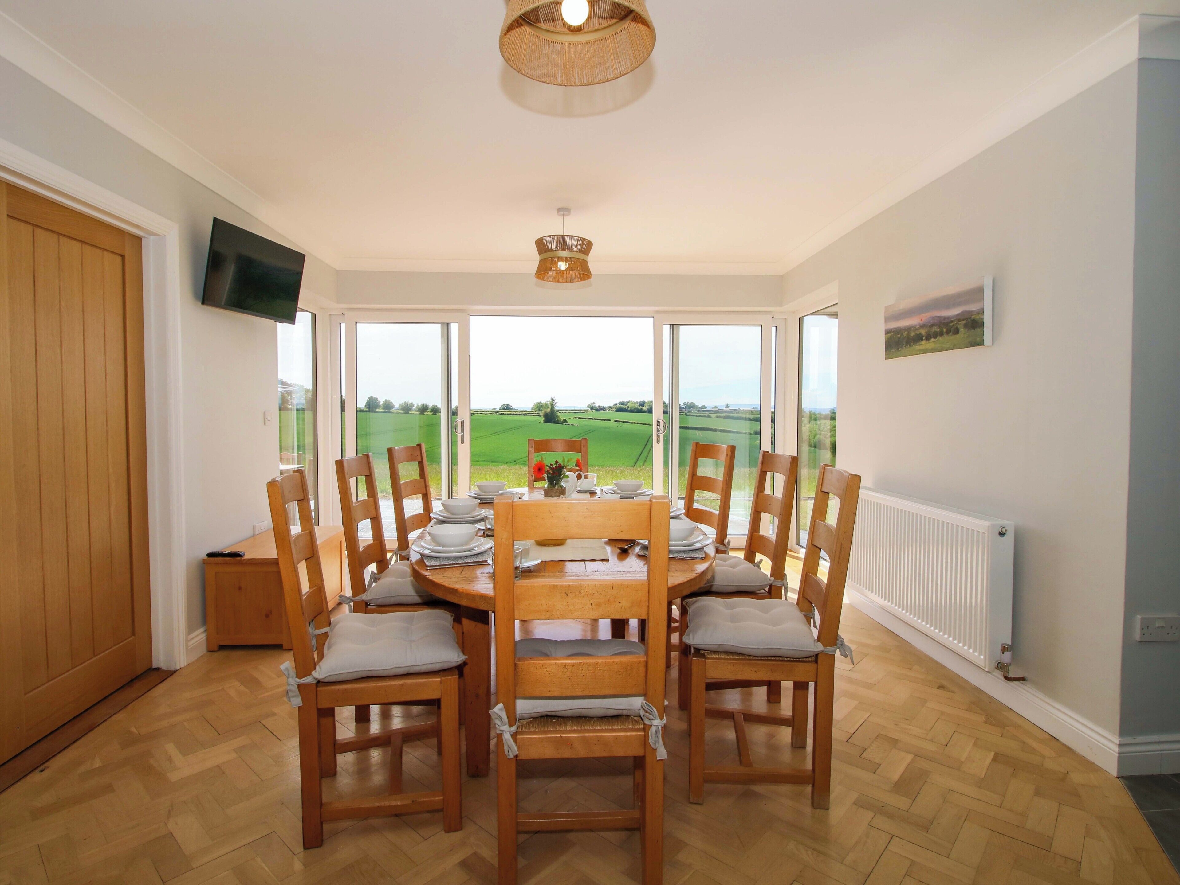 Un coin repas avec une table dressée pour six personnes, entouré de chaises en bois dotées de coussins. Un téléviseur est fixé au mur, et une grande fenêtre offre une vue sur un paysage verdoyant.