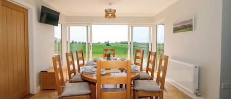A dining area with a table set for six, surrounded by wooden chairs with cushions. A television is mounted on the wall, and a large window offers a view of a green landscape.
