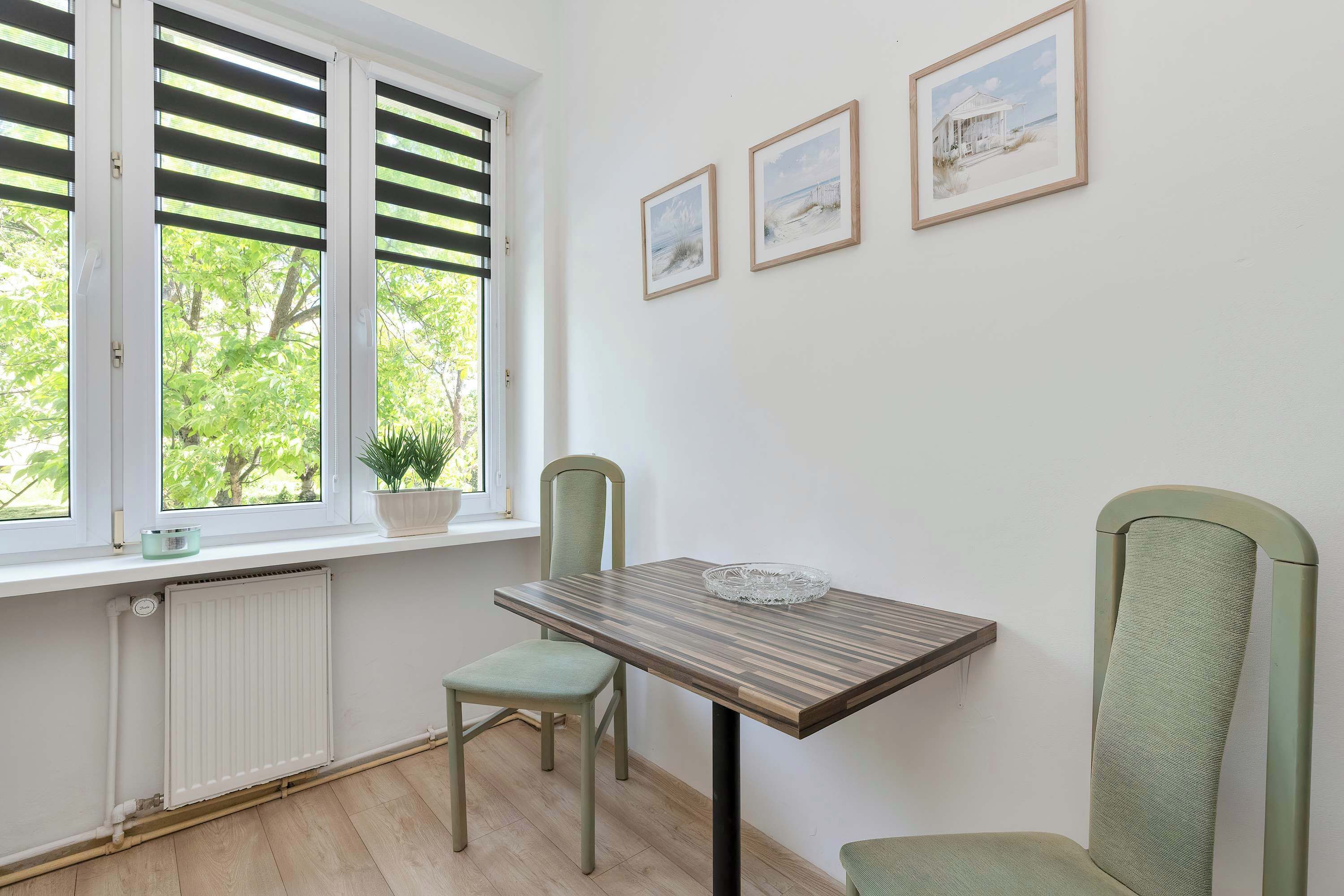 Un coin repas comprenant une table, des chaises, une fenêtre avec des stores, un radiateur et des tableaux encadrés accrochés au mur.