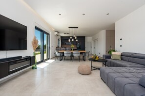 Ein modernes Wohnzimmer mit Flachbildfernseher, einer Couch, einem Essbereich mit Stühlen und einer Küche im Hintergrund.
