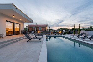 Ein moderner Poolbereich im Freien mit Liegestühlen, einem klaren Pool und einem Gebäude mit roter Ziegeldach.