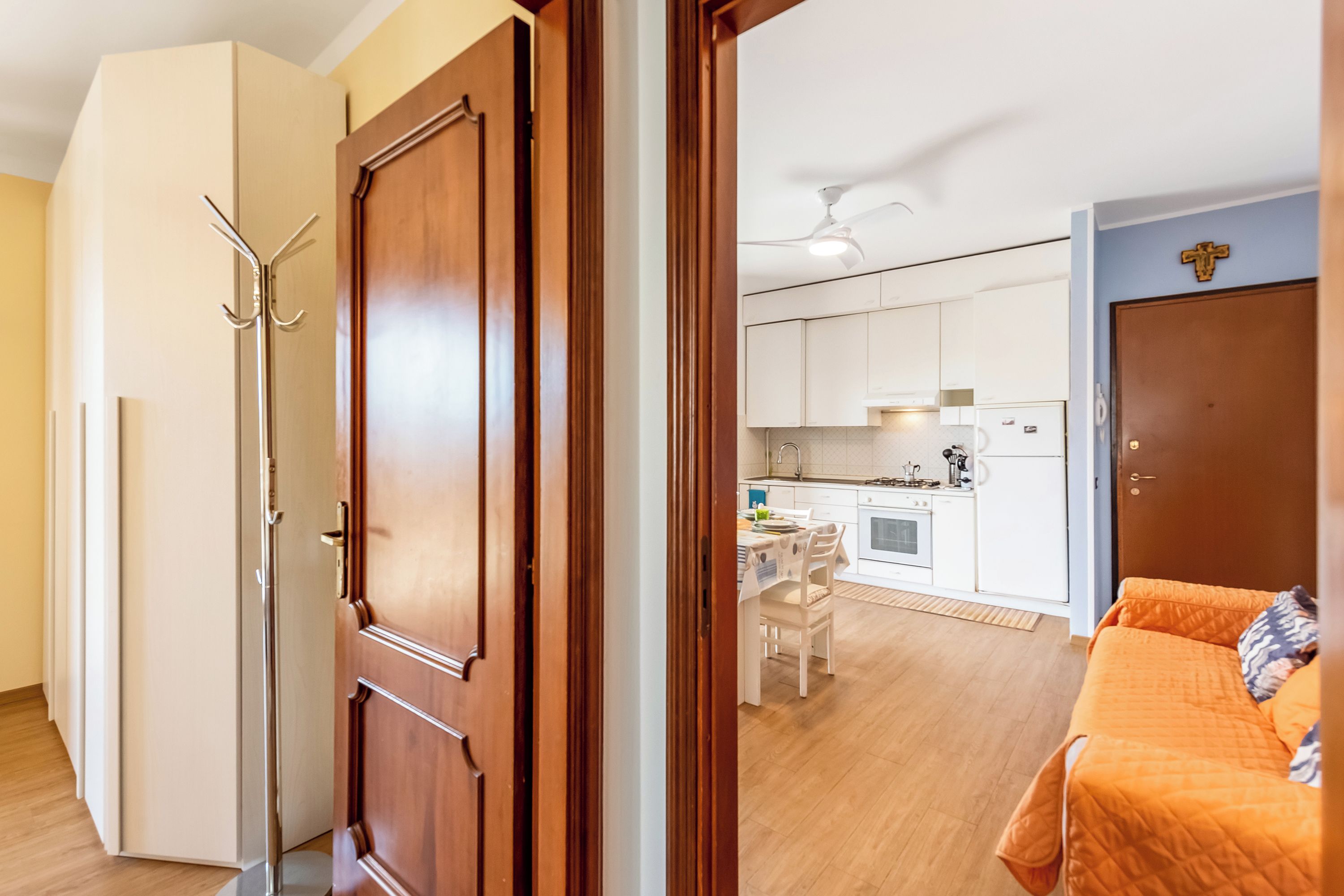 A room with a wooden door, a bed with an orange cover, a kitchen area with white cabinets, and a dining table with chairs.
