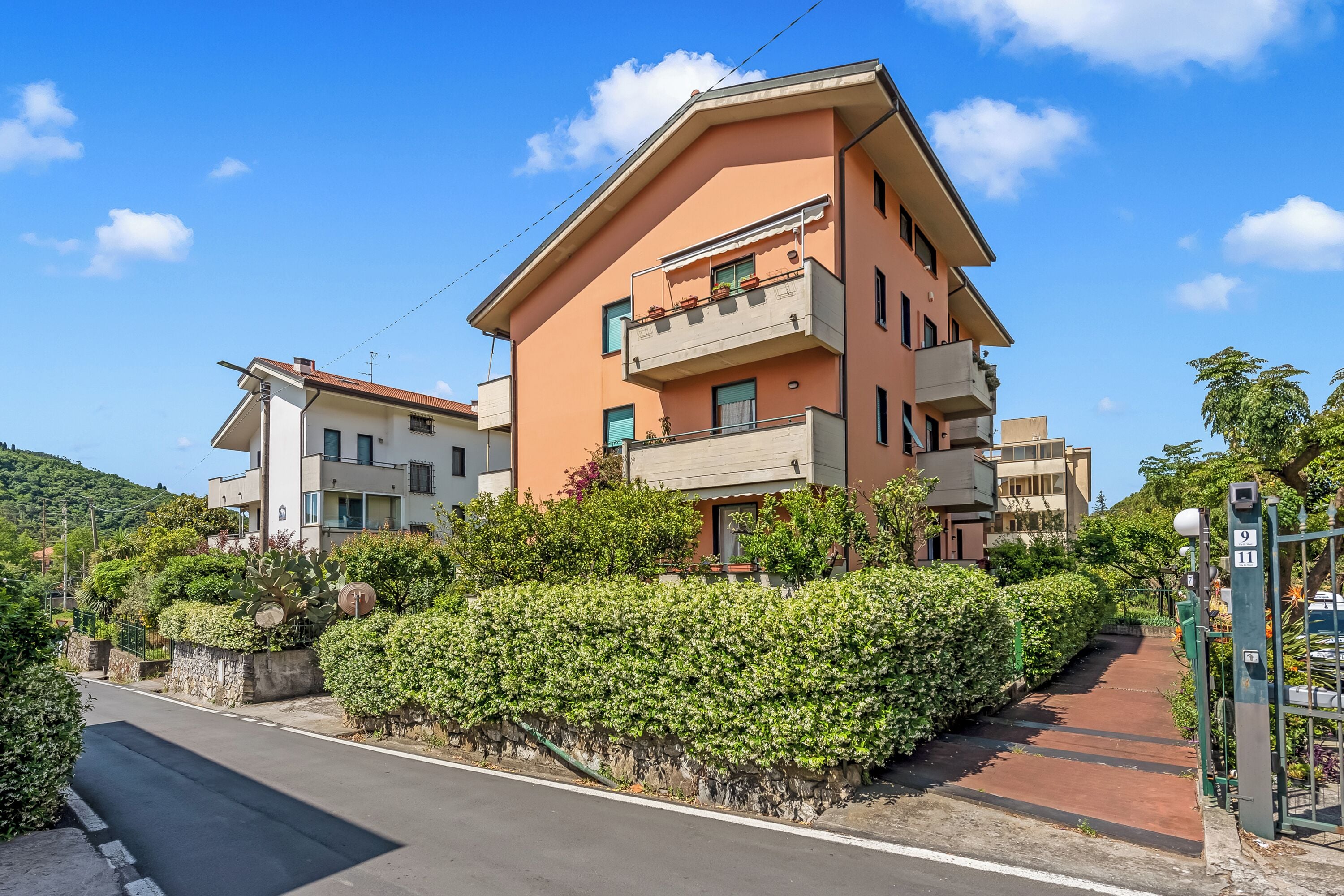 A multi-story residential building with balconies, a paved driveway, and a green hedge.