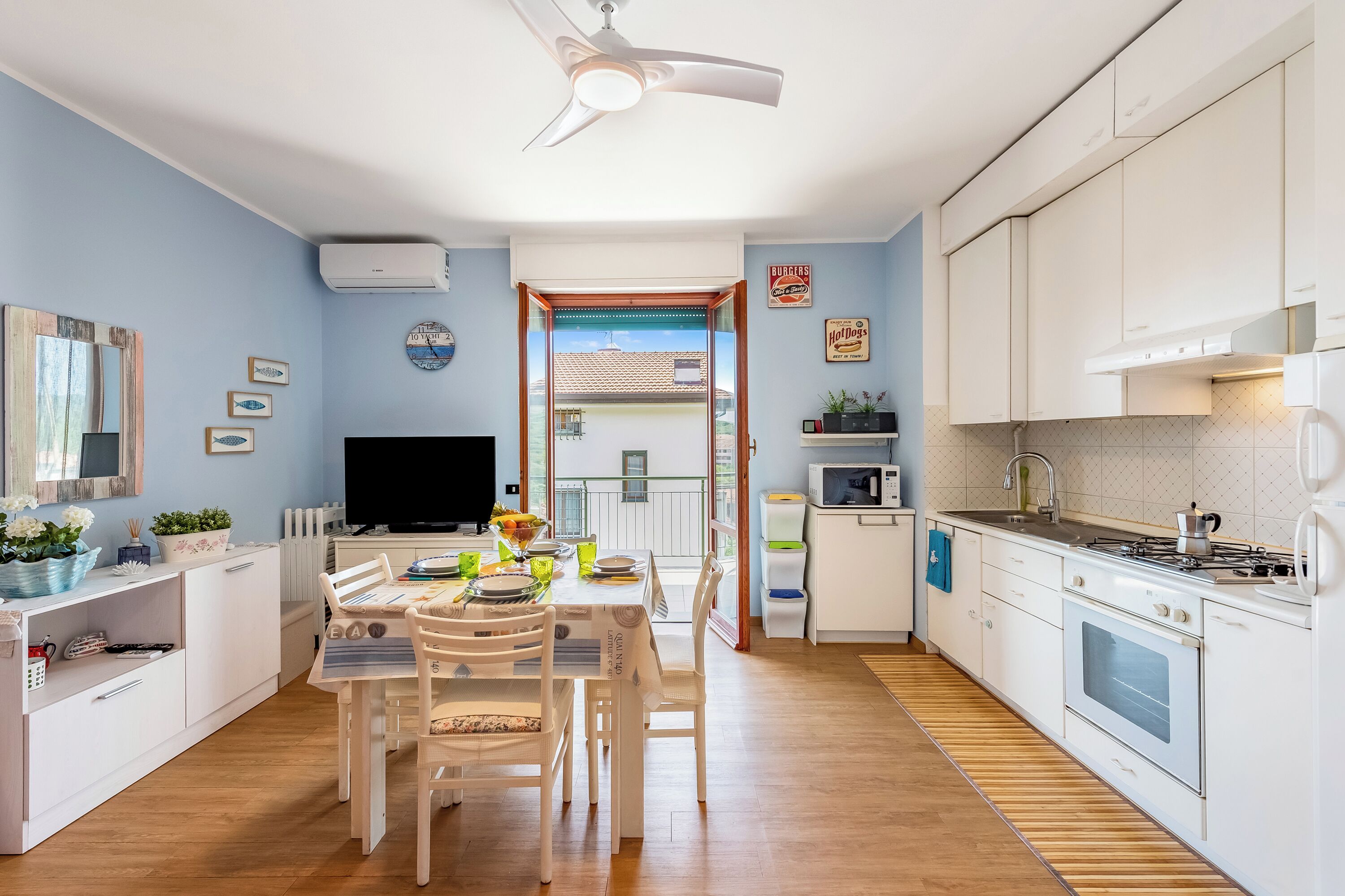 Una cocina moderna con armarios blancos, un comedor con mesa y sillas, un televisor y vistas al exterior a través de un ventanal.