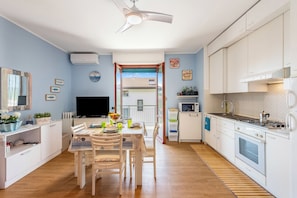 Eine moderne Küche mit weißen Schränken, einem Essbereich mit Tisch und Stühlen, einem Fernseher und Blick durch eine Glastür nach draußen.