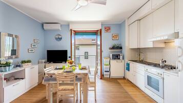 Una cocina moderna con armarios blancos, un comedor con mesa y sillas, un televisor y vistas al exterior a través de un ventanal.