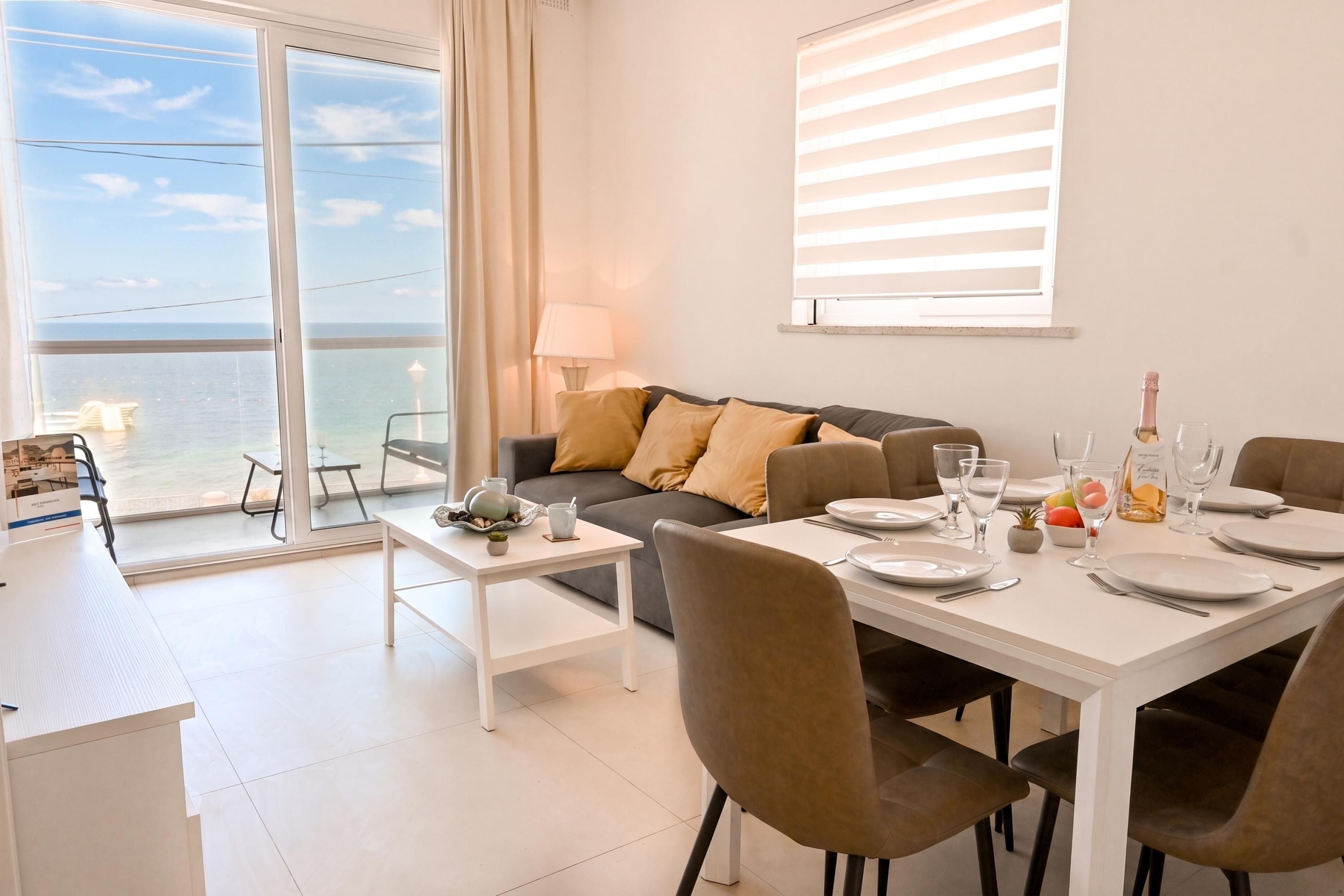 A modern living room with a balcony overlooking the sea, a dining area with a table set for two, and a sofa with cushions.