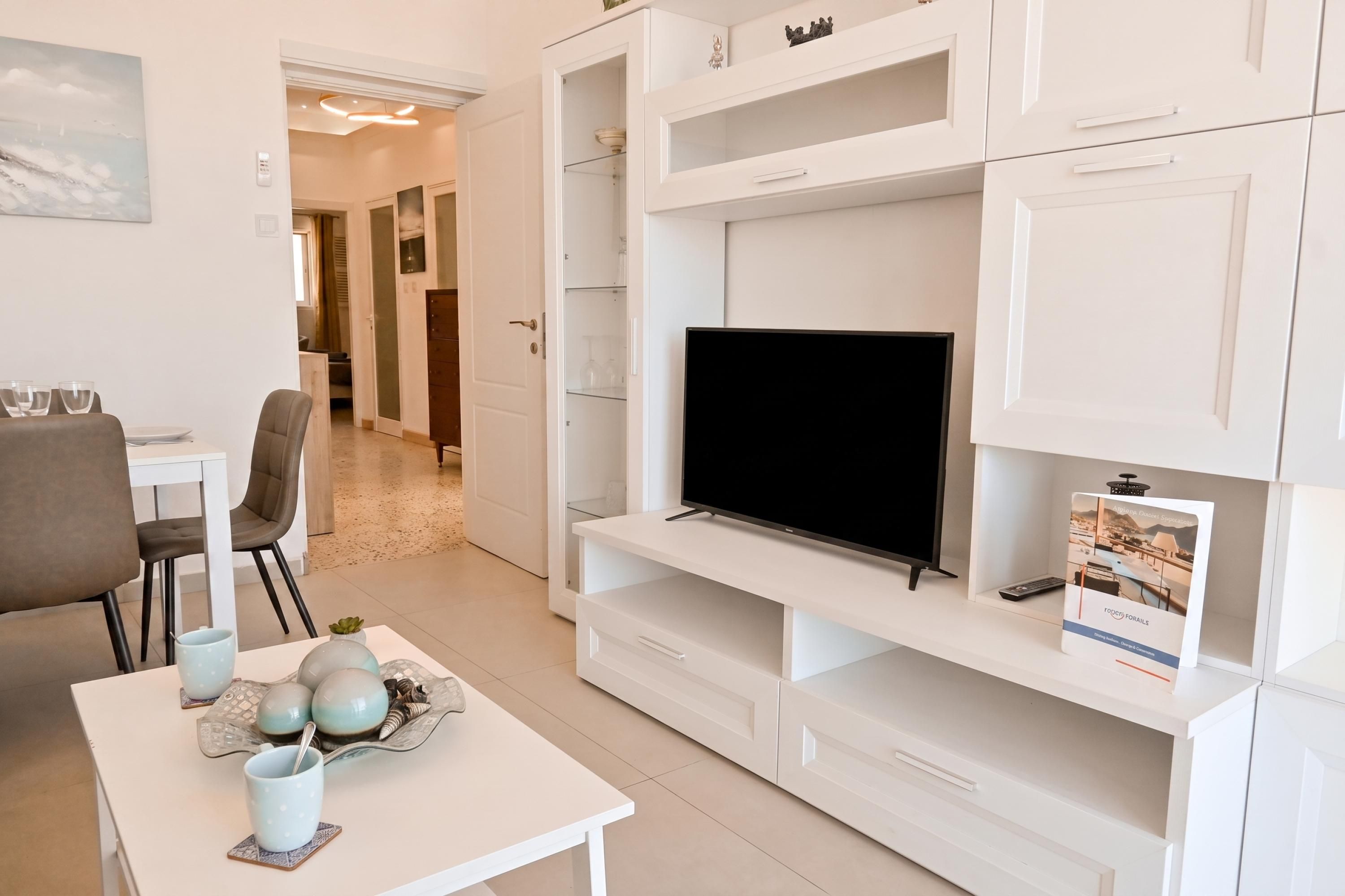 A modern living room with a flat-screen TV, white cabinetry, a dining area with a table and chairs, and a hallway leading to another room.