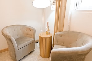 Zwei gepolsterte Sessel, ein Holztisch mit Flasche und Glas sowie eine Stehlampe in einem Raum mit Fenster und Vorhängen.