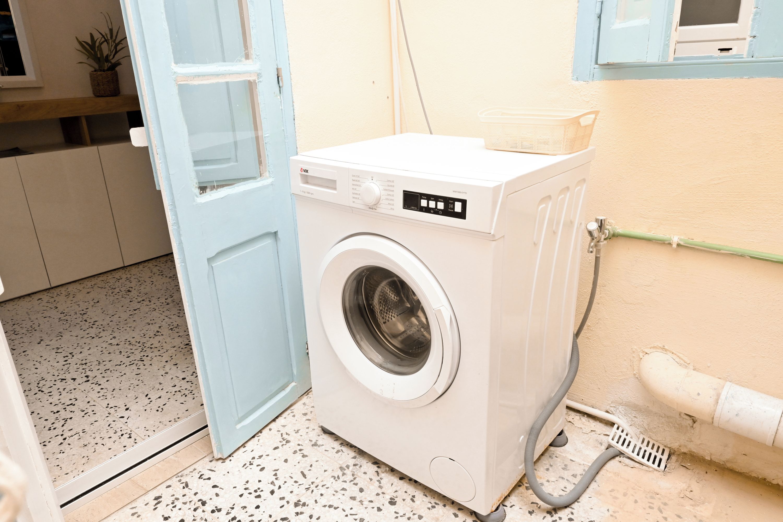 A compact, white front-loading washing machine in a small laundry room with a tiled floor and a blue door.
