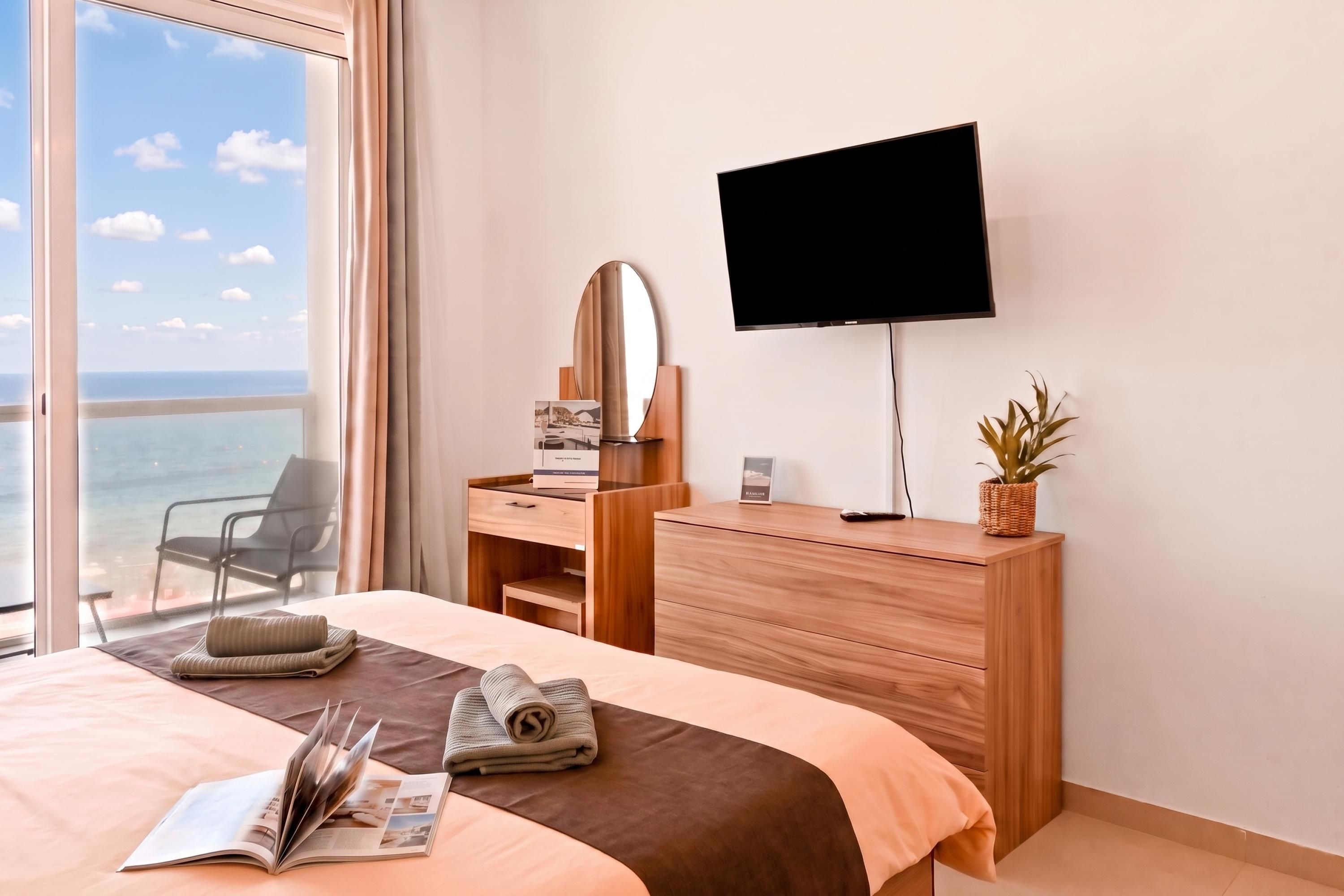 A hotel room with a bed, a desk with a flat-screen TV, a chair, a window with a view of the sea, and a potted plant.