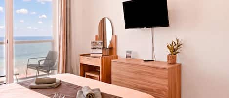 A hotel room with a bed, a desk with a flat-screen TV, a chair, a window with a view of the sea, and a potted plant.