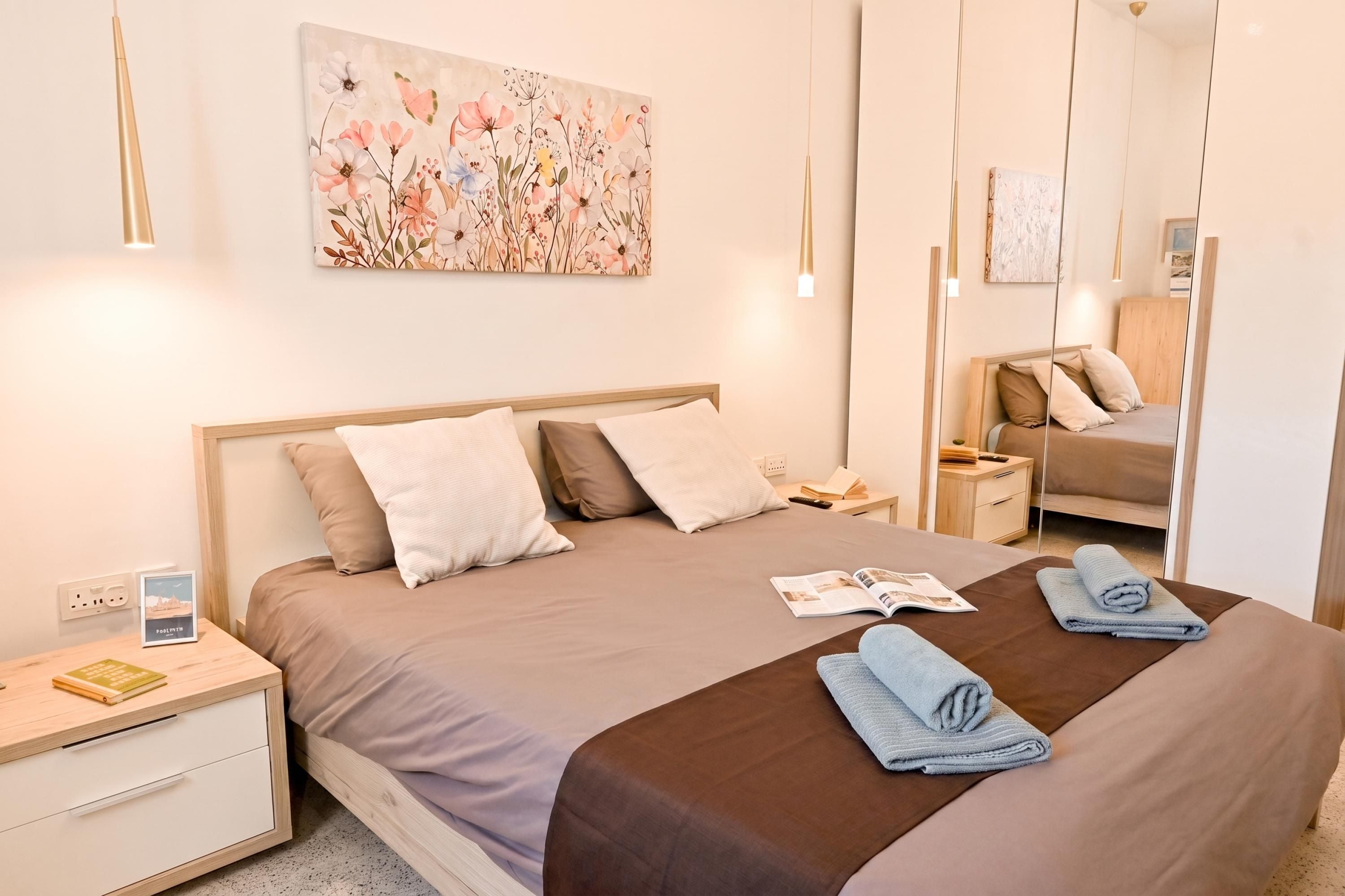 A modern bedroom with a large bed, bedside tables, a nightstand, a mirror, and a floral artwork on the wall.