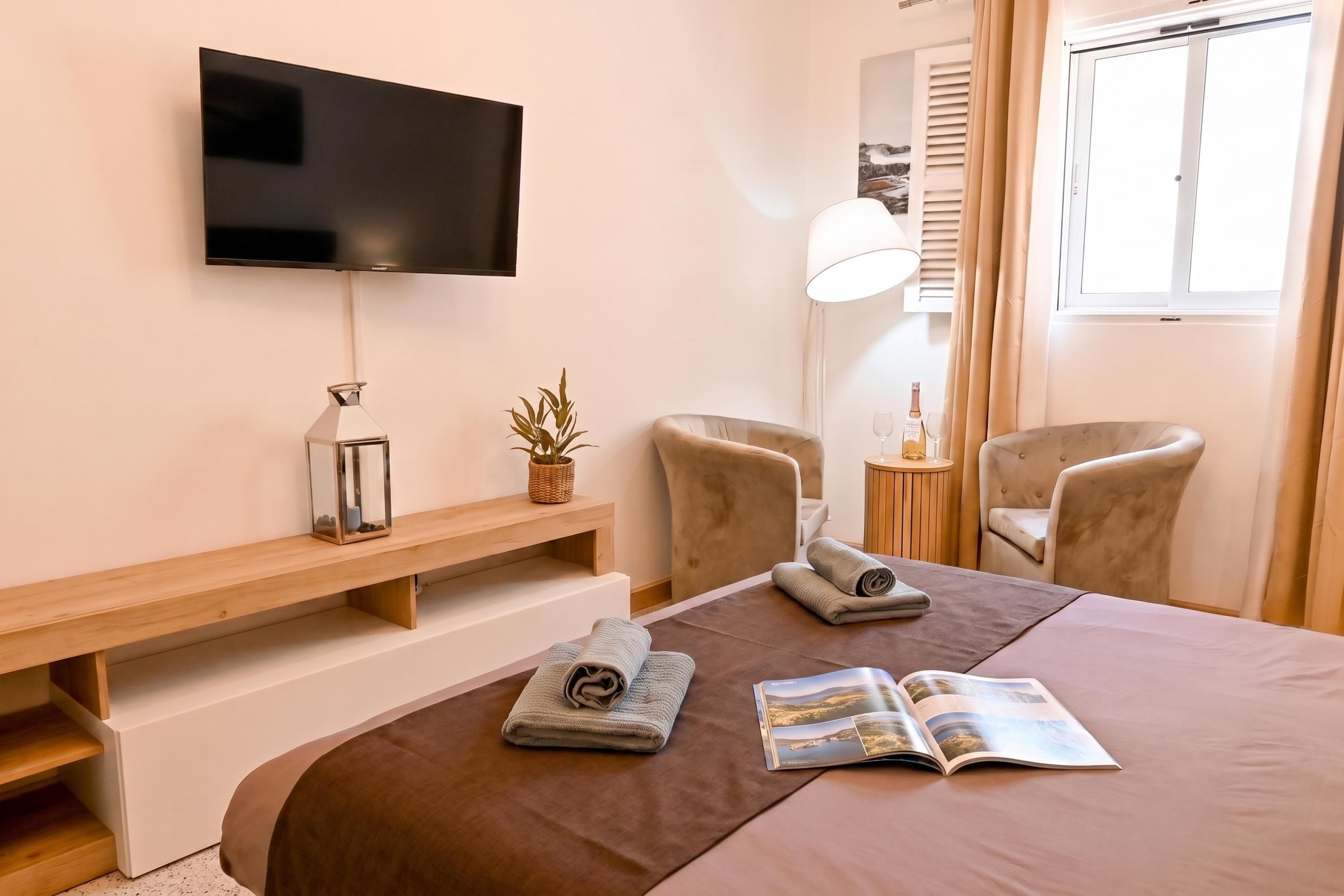 A hotel room with a flat-screen TV, a wooden bench, two chairs, a bedside table with a lamp, a window with curtains, and a bed with towels and a magazine.