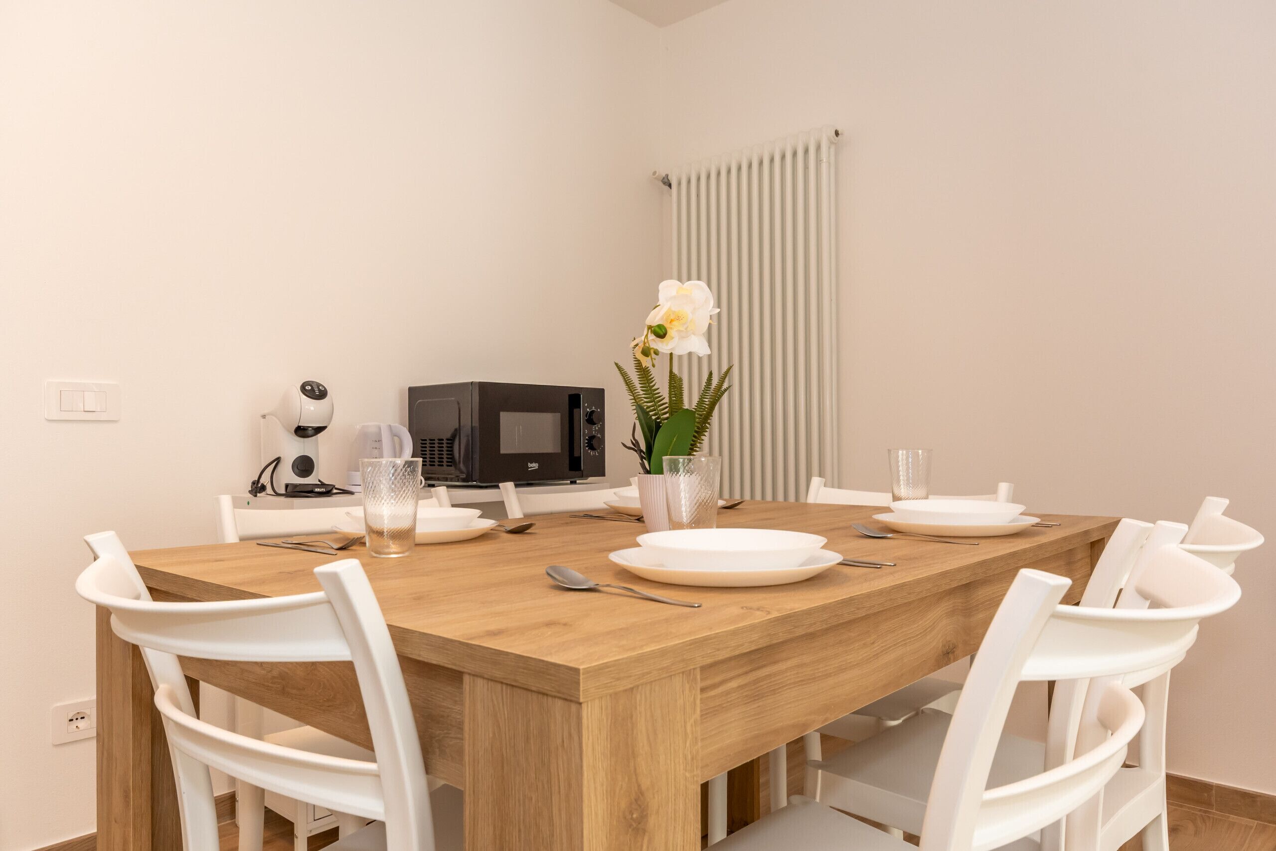 A dining area with a wooden table, white chairs, a microwave, a small potted plant, and a white radiator.