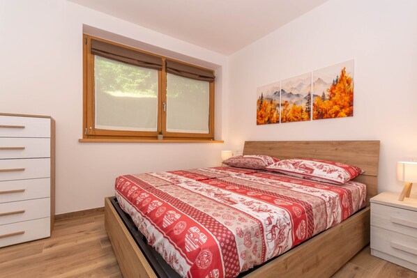 A bedroom with a bed, a dresser, a nightstand, a window with blinds, and a painting on the wall. - Alpine Haven - Deer Lodge (Cencenighe Agordino)