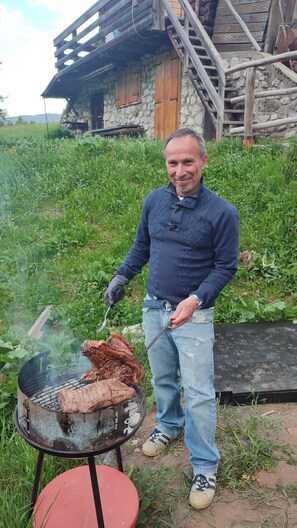 Ein Mann grillt Fleisch im Freien, im Hintergrund ein rustikales Haus.