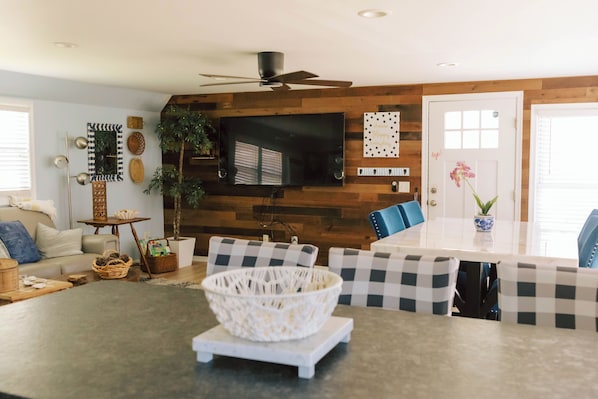 A modern living room with a dining area, a flat-screen TV mounted on a wooden wall, a ceiling fan, and a door with a decorative sign.