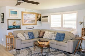 A living room with a beige sectional sofa, a wooden coffee table, a patterned rug, and wall art including a ship painting and a beach sign.