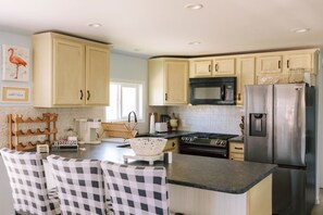 A kitchen with light-colored cabinets, a stainless steel refrigerator, a microwave, and a stove. There are chairs with checkered upholstery and a welcome sign.