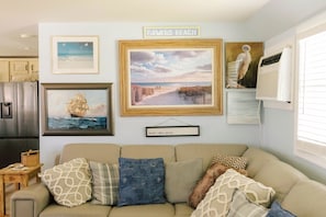 A living room with a beige sofa, framed artwork, a sign reading 'Bowers Beach,' and a kitchen in the background.