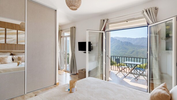 A bedroom with a bed, a TV, and a balcony with a view of mountains.