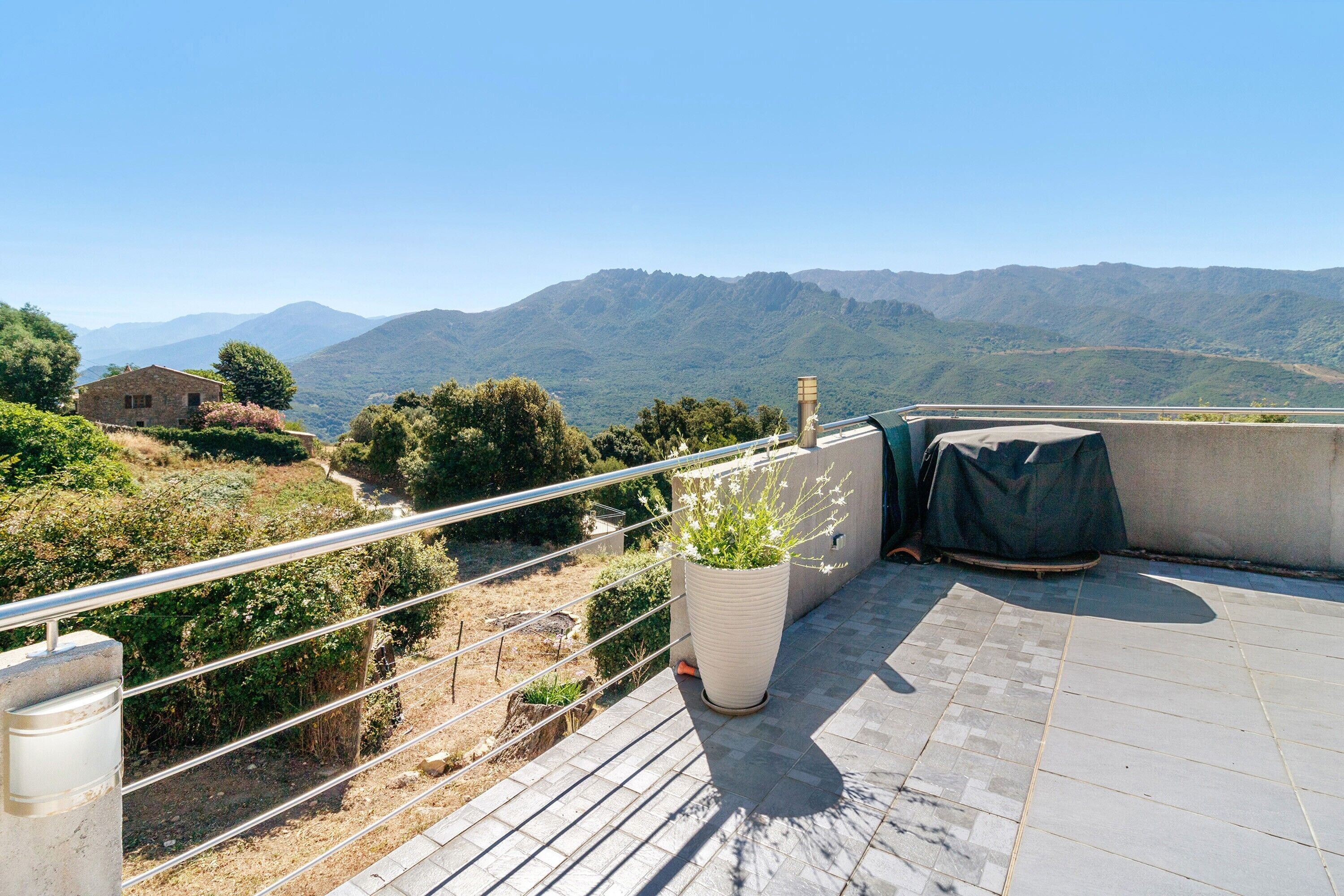 A rooftop terrace with a view of mountains, a potted plant, and a covered grill.