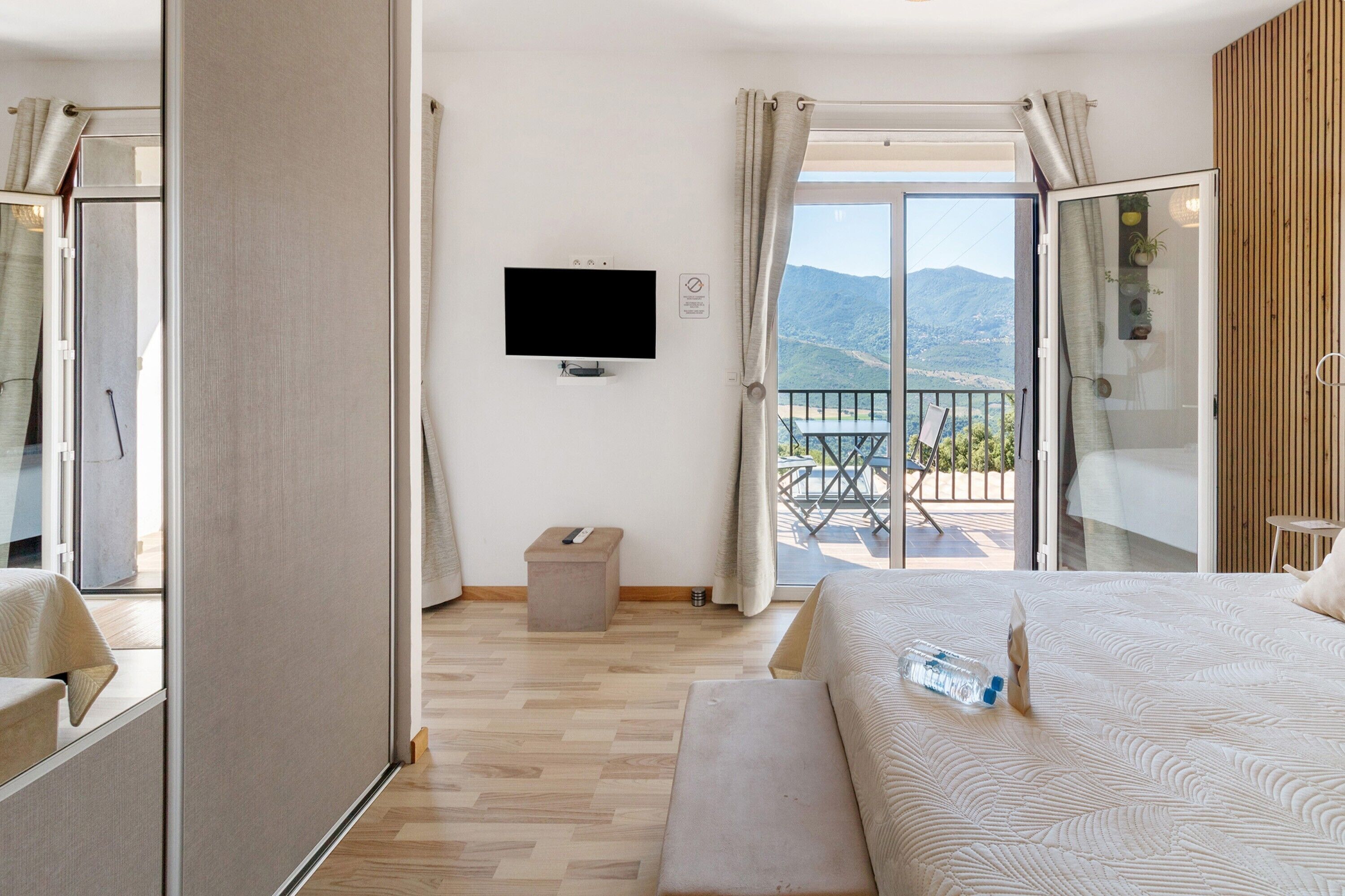 A hotel room with a bed, a television, a small table, a bench, and a balcony with a view of the mountains.