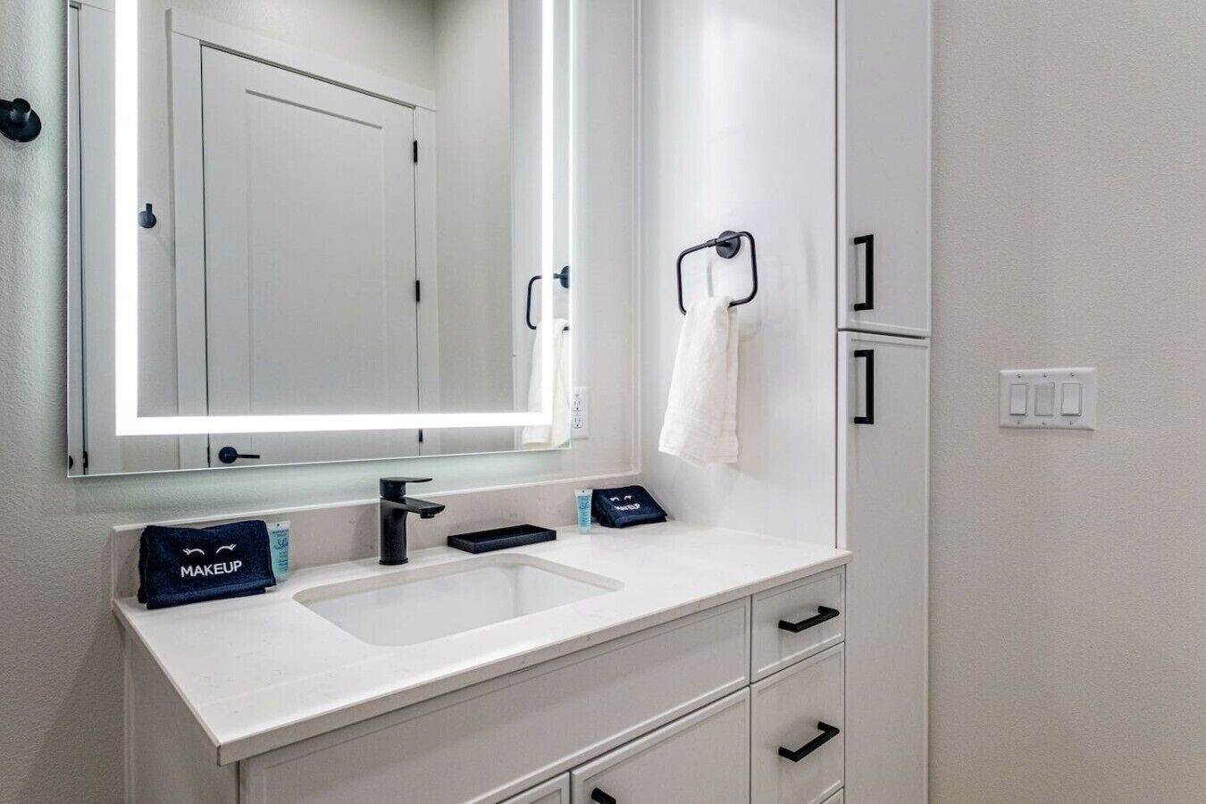A modern bathroom with a white sink, black faucet, and a large mirror with built-in lighting. There is a towel rack with a white towel and a makeup bag on the counter.