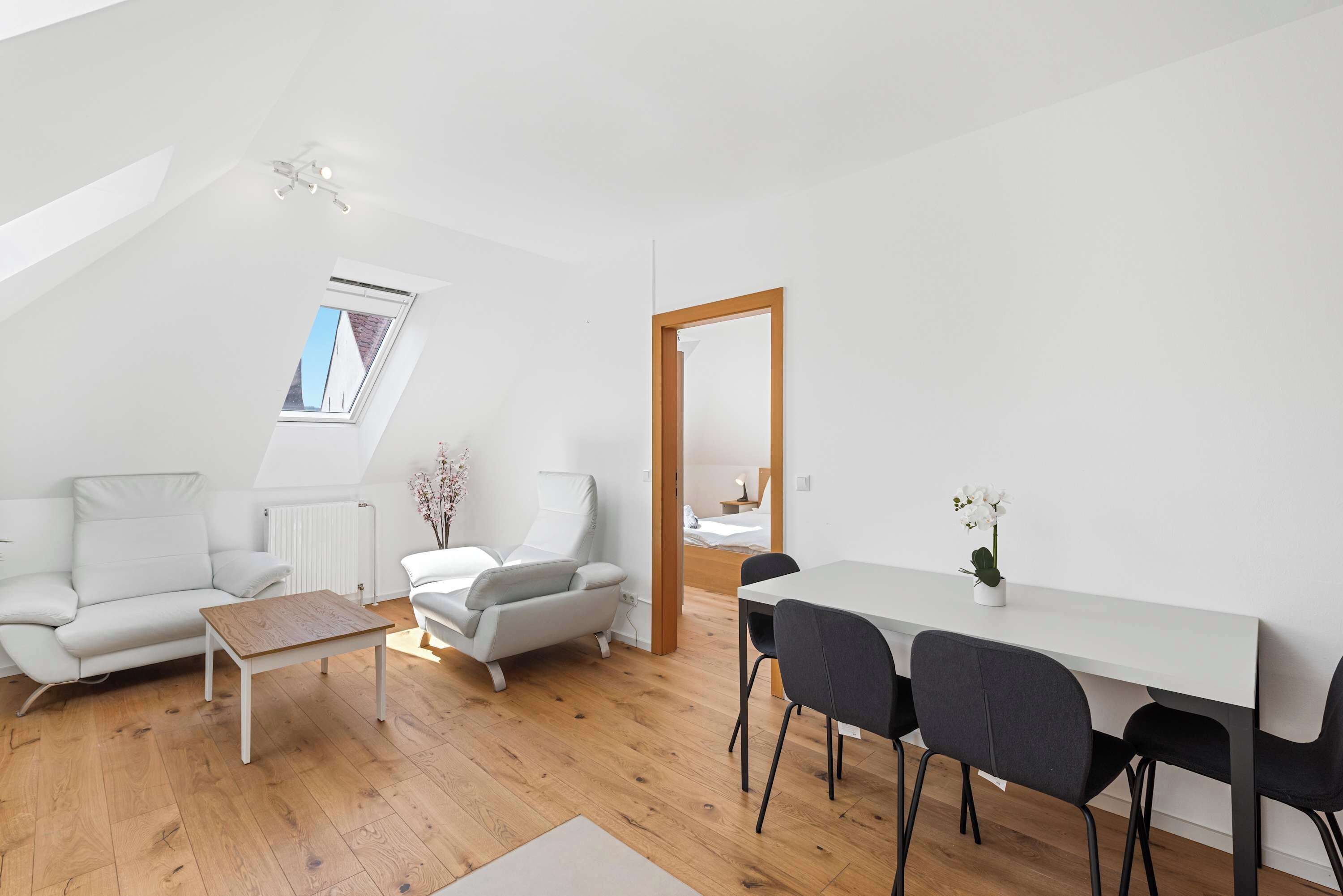 Un appartement moderne de style « loft » avec un plafond en pente, un coin repas comprenant une table et des chaises blanches, un canapé blanc et une table basse en bois.