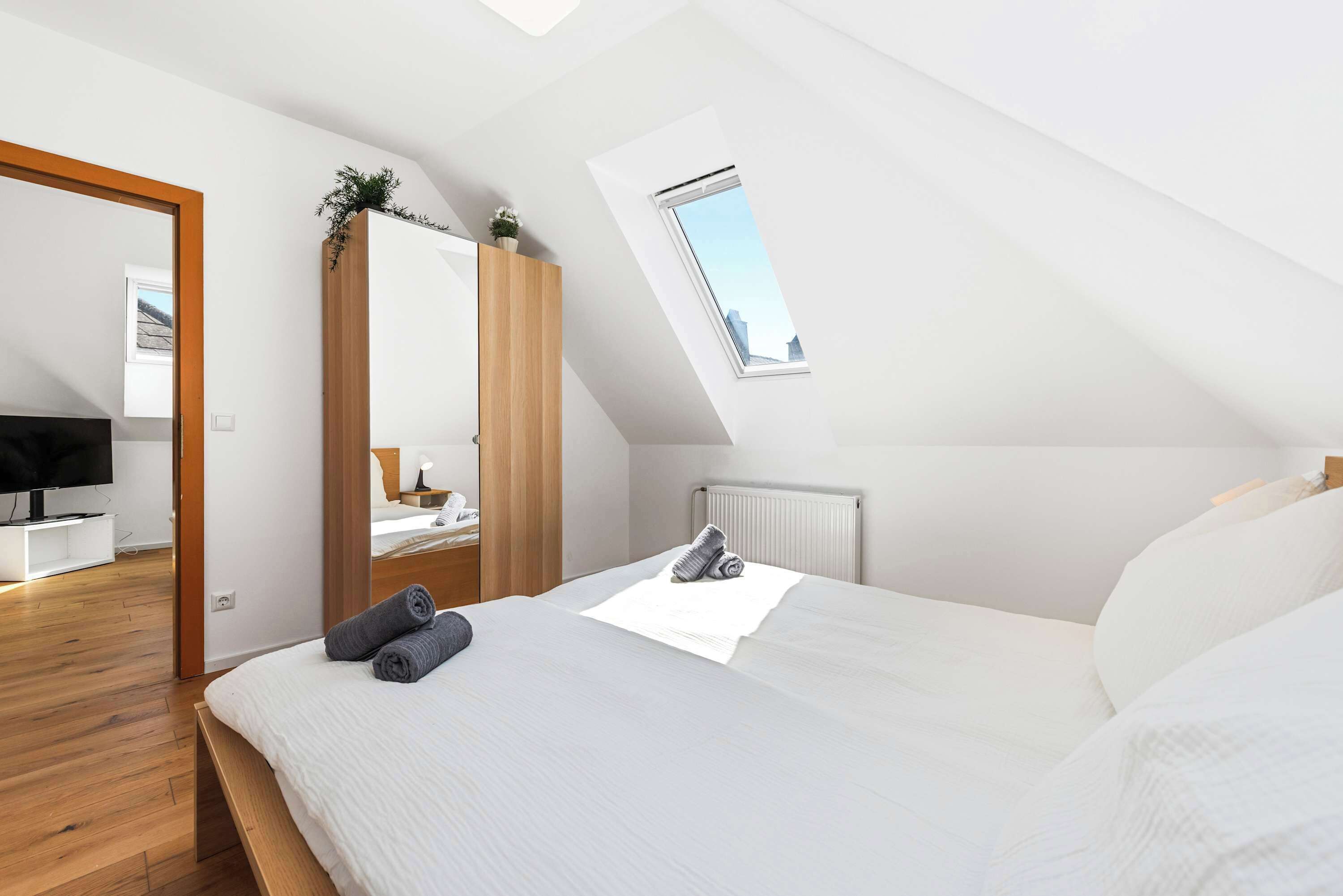 A bedroom with a sloped ceiling, a skylight, a bed with pillows, a TV, and a wooden wardrobe.