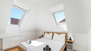Un dormitorio con techo inclinado, tragaluces, una cama con ropa de cama blanca, una mesita de noche con lámpara y un radiador.