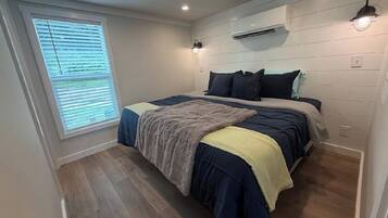 A bedroom with a bed, a window with blinds, an air conditioning unit, and a wall-mounted light fixture.