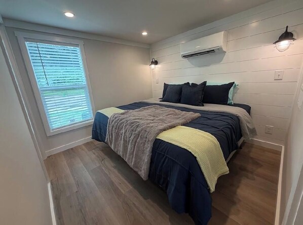 Un dormitorio con cama, ventana con persianas, unidad de aire acondicionado y una lámpara fijada a la pared.