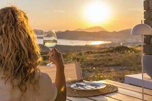 A person sitting at a table with a glass of white wine, overlooking a scenic view of a body of water and mountains during sunset. - Villa Nabia II, Seaview and Sunset view, Private Pool (Adamas)