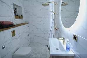 Un baño con piso de mármol blanco, inodoro blanco, lavamanos blanco con grifo dorado y ducha con cabezal dorado.