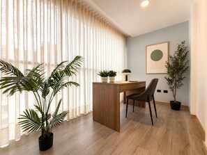 Una habitación moderna con un escritorio de madera, una silla, una planta en maceta y un ventanal con cortinas translúcidas.