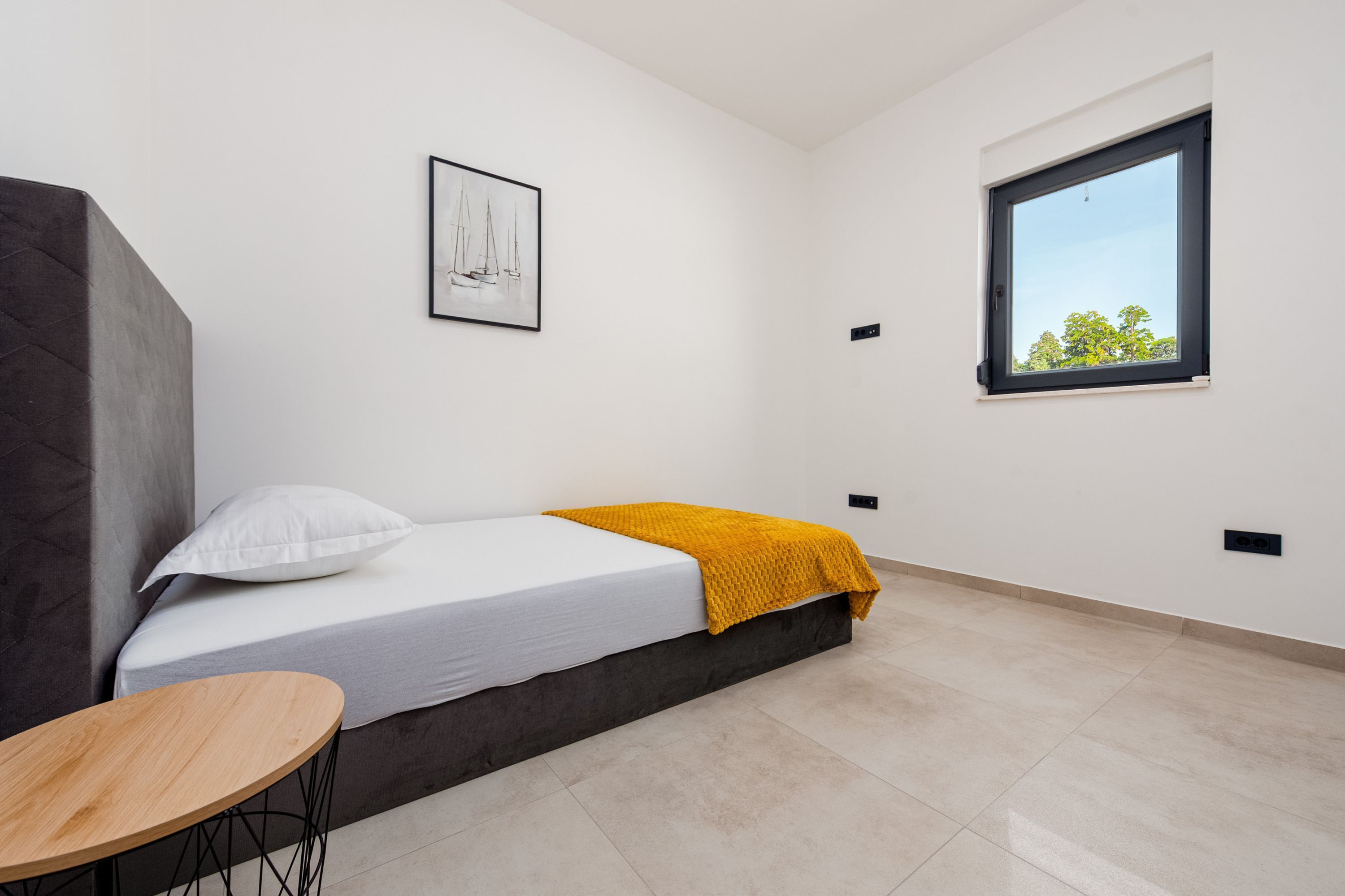 A minimalist bedroom with a bed, a single window, a small table, and a framed picture on the wall.