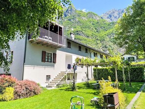 Ein zweigeschossiges Gebäude mit einem Balkon, einer Treppe zum Eingang und einem Garten mit verschiedenen Pflanzen.