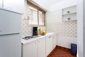 Eine kleine Küche mit weißen Schränken, einem weißen Kühlschrank, einem Fenster mit Jalousien, einem Spülbecken, einem Herd und einem kleinen Regal mit Pflanzen.