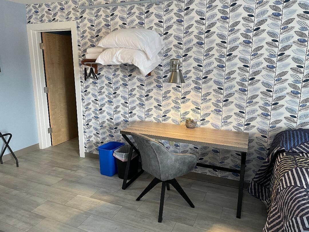 A small room with a wooden desk, a grey chair, a blue trash bin, and a patterned wall.
