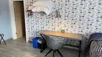 A small room with a wooden desk, a grey chair, a blue trash bin, and a patterned wall.